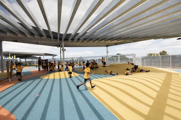An expansive covered outdoor recreation area with children participating in sports and play activities on a brightly coloured rubberised surface. A large exposed-beam canopy creates strong linear shadows across the blue, yellow, and orange flooring, with open sides, fencing, and additional activity equipment visible beyond.