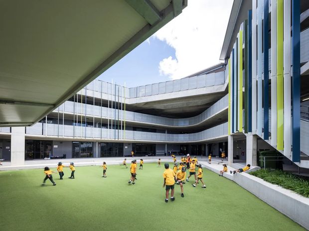 A contemporary multi-storey institutional courtyard viewed from an elevated covered walkway. The central landscaped synthetic-grass recreation area is occupied by children in sports uniforms, surrounded by curved concrete balconies, extensive glazing, metal balustrades, and colourful vertical facade fins in blue, white, and lime green.