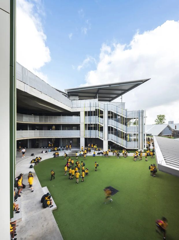 A contemporary multi-level school courtyard viewed from an elevated position. The central outdoor space has artificial green turf, concrete seating edges, and many students in sports uniforms. Surrounding buildings feature exposed circulation stairs, perforated metal balustrades, extensive concrete construction, and a large projecting metal canopy. A pitched-roof school building is visible beyond the courtyard.