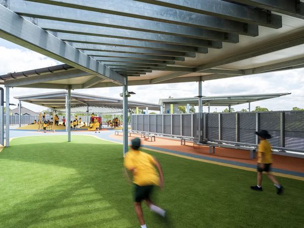 A covered outdoor recreation area with extensive steel-framed shade structures and translucent roof panels. Artificial green turf occupies the foreground, with children in yellow sports clothing, benches along a dark privacy fence, and colourful play or activity courts in the background. The space is bright and functional, with a strong emphasis on sheltered outdoor use.