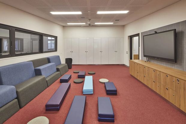 A spacious contemporary media or multipurpose room with rows of low modular upholstered seating and floor cushions arranged around a large wall-mounted screen. The room has a red carpeted floor, pale walls, extensive dark-framed interior glazing, full-height white storage cupboards, and a timber drawer console beneath a dark textured feature wall. Fluorescent ceiling panels and acoustic perforated ceiling tiles give the space a practical institutional or commercial character.