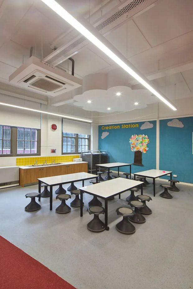 A bright, colourful institutional dining and activity room, likely a school cafeteria or creative learning space. Multiple rectangular tables with fixed stools are arranged across a polished light-grey floor. A timber-fronted kitchenette with a vivid yellow tiled splashback runs along the left wall, while the rear wall features a blue mural reading “Creation Station,” with cloud and tree artwork. The room has large windows with blinds, exposed services, suspended linear LED lighting, and a sculptural cloud-shaped ceiling feature.