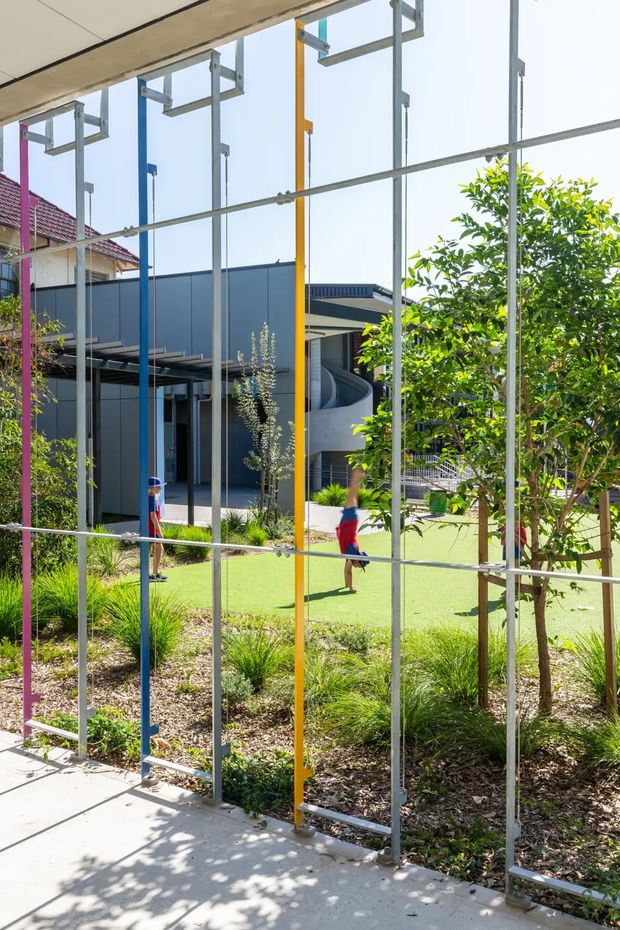 A bright exterior view through a colourful metal structural frame into a landscaped courtyard or school-like outdoor area. The scene includes a contemporary dark-clad building with large glazing, a covered walkway, lawn, young trees, ornamental grasses and two people in the distance. The foreground shows a concrete path and a prominent grid of exposed silver framing with pink, blue and yellow vertical posts.