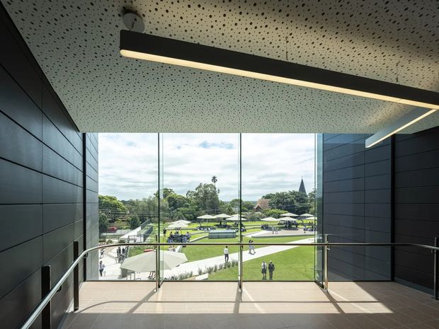 A modern institutional interior landing or corridor overlooks a landscaped courtyard through full-height floor-to-ceiling glazing. The space has dark horizontal wall panels, a perforated acoustic ceiling, a long linear pendant light, tiled flooring, and sleek metal-and-glass balustrades. Outside are lawns, umbrellas, people, and a distant church spire.