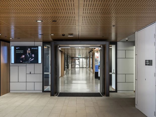 A contemporary institutional or commercial building entry and hallway. The image shows a wide tiled foyer leading through dark-framed glazed doors into a long bright corridor. The interior features a perforated or slatted timber ceiling, light tiled flooring, pale segmented wall panels, dark metal and glass door frames, integrated display screens, and white service or storage doors. The space is clean, functional, and moderately bright.