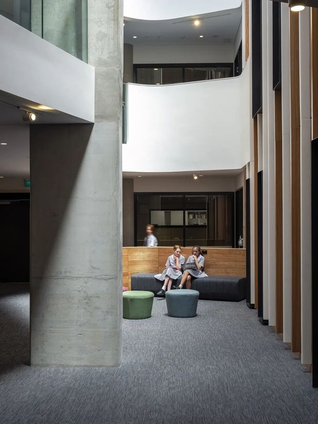 A contemporary multi-level interior atrium or lobby with a double-height void, curved white balcony edges, exposed concrete columns, dark-framed glazing, and timber-lined built-in seating. Grey carpet covers the floor, with a dark upholstered bench and small green and blue ottomans. Children are seated in the lounge area, while vertical timber and dark panels line the right-hand wall. Recessed and spot lighting creates a calm, refined institutional or commercial atmosphere.