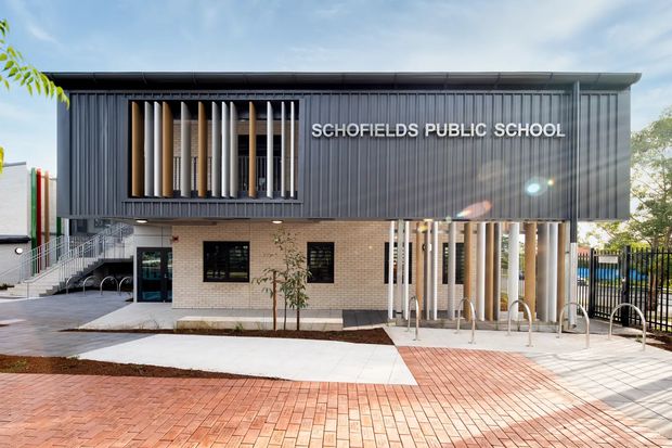 Exterior view of Schofields Public School, showing a contemporary two-storey institutional building with dark vertically ribbed metal cladding, a light brick lower level, projecting sun-shading fins, broad glazing, accessible stairs and ramps, bicycle hoops, and a paved brick forecourt.