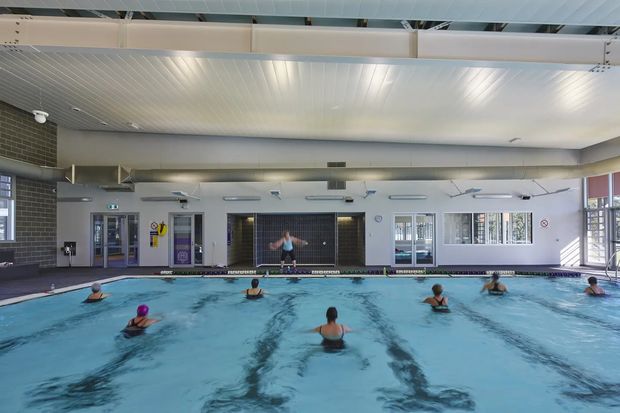 Indoor swimming pool in a spacious aquatic recreation facility, with multiple marked pool lanes, swimmers participating in a class, a tiled feature wall, extensive glazing, and a white ribbed ceiling with exposed structural elements.