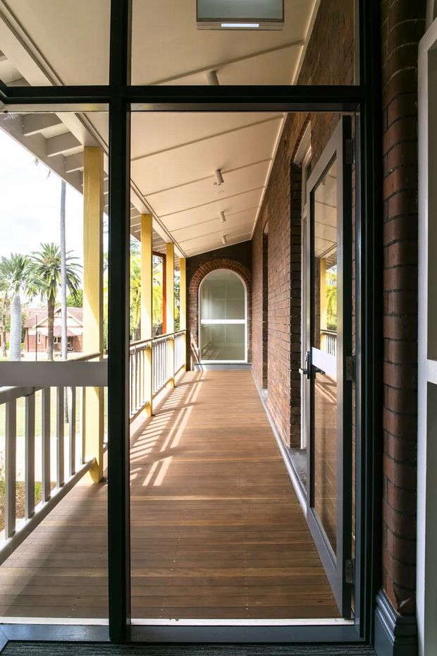 View from inside through a black-framed glazed door onto a long covered balcony or verandah. The outdoor passage has timber decking, a white-lined ceiling with recessed and surface-mounted lights, brick walls with arched window detailing, and a simple balustrade. Tropical palms and neighbouring residential buildings are visible beyond the railing. The image emphasizes the sheltered exterior circulation space and connection between interior and outdoors.