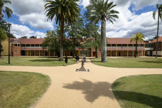 A broad landscaped institutional courtyard with curved pale gravel pathways, lawns, mature palm trees, and a central pedestal sculpture. A symmetrical two-storey brick building with covered walkways, repetitive windows, and red pitched roofs forms the backdrop under a partly cloudy sky.
