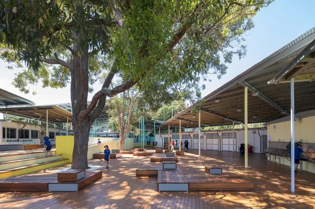 A bright outdoor courtyard or campus space with extensive timber decking, bench seating built around a mature tree, and covered open-sided structures with corrugated metal roofs. Low-scale pavilion-like buildings, colourful walls, playground or recreational elements, and several people are visible. The scene has a functional, communal architectural character with abundant shade and greenery.