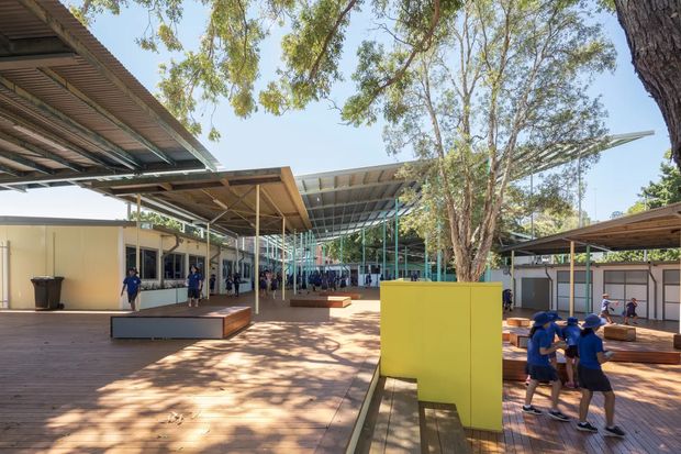 A spacious outdoor school or community courtyard with extensive steel-framed covered walkways and timber decking. Low modular buildings surround the open space, with large established trees providing shade. Bright yellow and turquoise architectural elements add colour, while students gather and move through the courtyard.