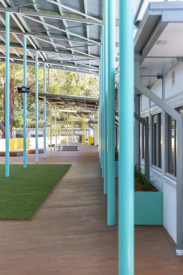 A bright covered outdoor walkway or courtyard beside a modern building, with a long timber-look deck, vivid turquoise cylindrical columns, exposed steel roof framing, planted built-in planters, and a strip of artificial grass. Yellow structural and play-like elements are visible farther along the sheltered space, with trees beyond.