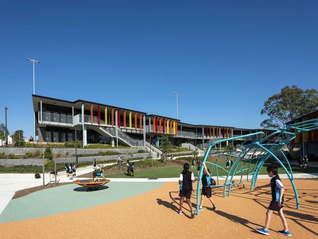 Exterior view of a contemporary multi-storey school building beside a colourful outdoor playground. The building has long horizontal wings, dark flat rooflines, extensive glazing, external stairs and balconies, and vertical panels in bright red, yellow and orange. The foreground contains turquoise climbing frames and orange and pale-green safety surfacing, with children using the play area.