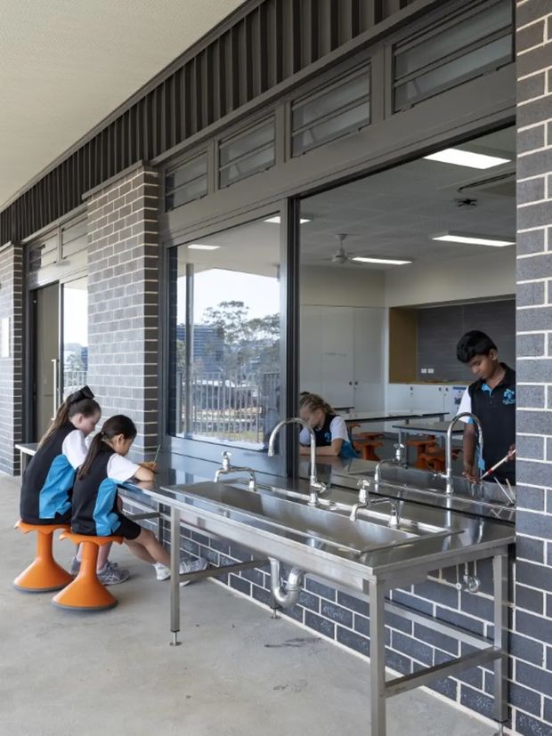 A covered outdoor learning and washing area beside a school building, with stainless-steel multi-basin sinks, taps, exposed plumbing, and children seated at orange stools. Dark grey brick walls, large glazed openings, high-level louvred ventilation panels, and a visible classroom interior define the space.
