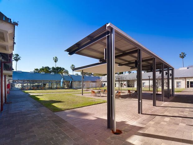 A sunny outdoor courtyard framed by low-rise buildings, with a large contemporary steel-and-timber pergola or covered pavilion over a paved seating area. The structure has slim dark metal posts, a flat roof with exposed timber soffit, and broad open sides. Brick-paved walkways and lawn surround the pavilion, with additional covered walkways and palm trees in the background.