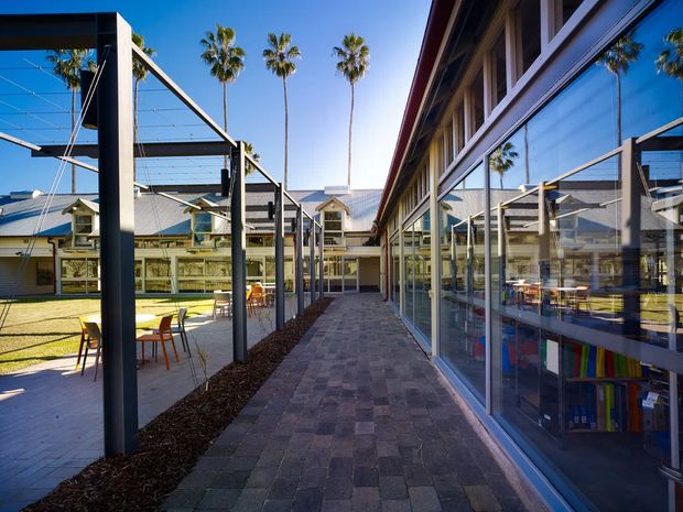 A sunlit paved courtyard walkway within a low-rise modern complex. Dark steel posts support open cable or wire shade structures over outdoor seating areas on the left, while a long glazed building facade with reflective windows and visible interior shelving runs along the right. Grey pitched metal roofs, pale walls, landscaped garden areas, and tall palm trees are visible beyond the courtyard. The composition emphasizes the exterior circulation space and strong reflections in the glazing.
