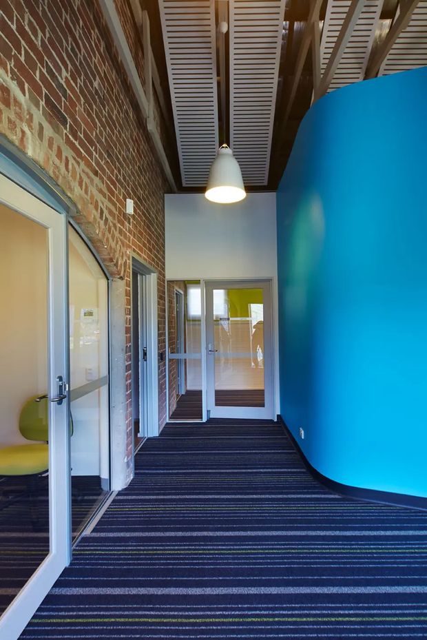 A narrow contemporary commercial corridor with exposed red brick walls, a vivid curved blue feature wall, glazed office doors, striped dark carpet, and an open slatted ceiling with visible structural beams. A simple white pendant light hangs centrally, and a small portion of seating is visible through the left doorway.