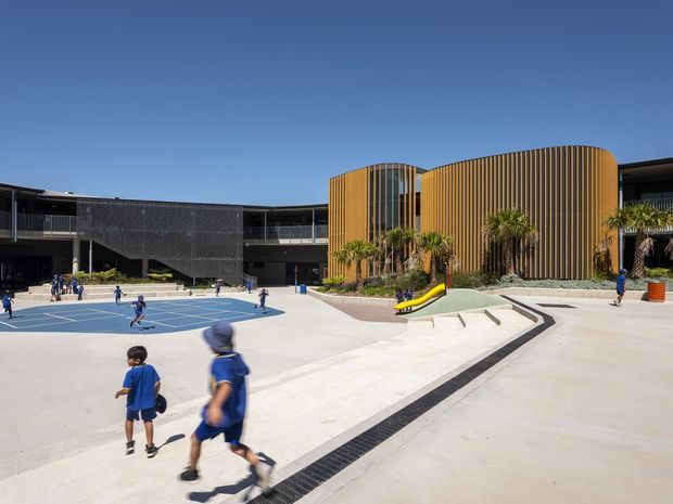 Bright daytime exterior view of a contemporary multi-storey school campus. The courtyard is dominated by a blue-lined sports court and broad pale concrete paving, with children using the space. A curved facade section features warm vertical timber-look battens and integrated planting, contrasting with darker perforated and glazed building wings. Accessible ramps, steps, linear drainage, and a small play slide are visible.