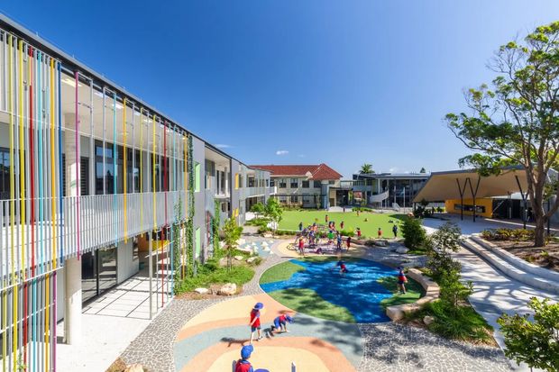 A bright aerial or elevated view of a colourful contemporary school or institutional campus arranged around a landscaped central courtyard. Multi-storey buildings feature extensive glazing, white façades, and prominent vertical sun-shading fins in varied colours. The courtyard includes lawns, play areas, patterned paving, large shade structures, and a small irregularly shaped blue water-play or pool feature. Children are visible using the outdoor recreation areas.