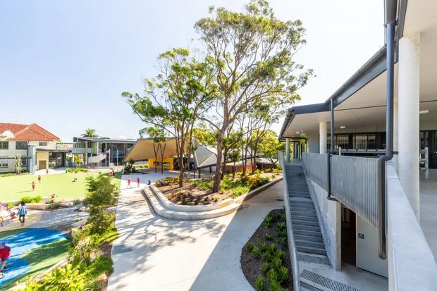 Bright outdoor view of a contemporary multi-level school or community campus, with broad concrete walkways, landscaped tree beds, covered verandas, stairs, and colourful recreational play areas. The buildings feature simple modern forms, extensive glazing, white rendered walls, and dark metal framing. The image is primarily an exterior circulation and landscape view.