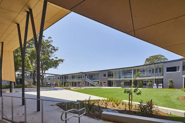 A contemporary multi-storey institutional or commercial building arranged around a landscaped courtyard. The view is framed by a broad timber-toned canopy with slender dark steel support columns, looking across paved paths, lawn, planted garden beds, and glazed circulation areas toward a long dark-clad building with repetitive windows and external stairs.