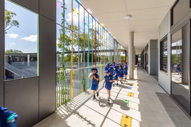 A bright, covered outdoor walkway in a contemporary school or institutional building. Children in blue uniforms walk along a wide concrete corridor beside full-height glazed and vertical metal screening, with classrooms or internal spaces opening along the right side. Strong sunlight creates striped shadows across the floor, and mature trees and an adjacent covered outdoor area are visible beyond the glazing.