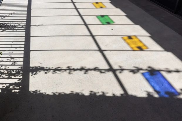 A close-up view of a paved outdoor surface divided into a regular grid of large pale rectangular panels, with small brightly coloured geometric markings in yellow, green and blue. Strong linear shadows from an overhead structure and foliage fall across the surface, creating a graphic, abstract composition.
