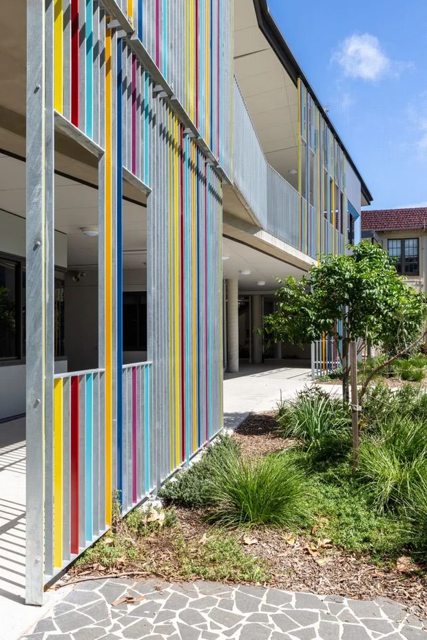 Exterior view of a contemporary multi-storey building with a highly distinctive facade of closely spaced vertical metal fins in bright yellow, red, blue, pink, and other colours. Open-air circulation balconies and shaded ground-level walkways frame a landscaped courtyard with ornamental grasses and a small tree. A patterned dark-and-light paved path occupies the foreground, while a neighbouring tiled-roof building is visible beyond.