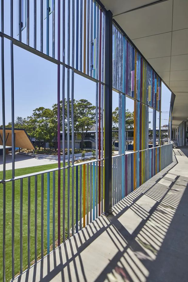 A bright exterior covered walkway beside a large glazed facade. Slim dark vertical framing and a colourful vertical screen of translucent or painted panels create strong striped shadows across the concrete path. Beyond the glazing is a landscaped institutional-looking courtyard with lawn, trees, and low buildings. The scene has a contemporary architectural character with a bold, playful facade treatment.