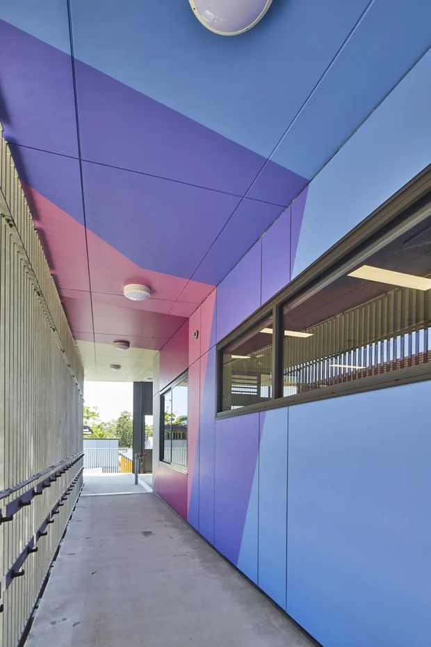 A covered exterior walkway beside a contemporary building, with a vivid geometric wall and soffit composed of blue, purple, and pink panels. The corridor has a smooth concrete floor, dark metal handrails, vertical metal screening on the left, and large glazed openings on the right revealing interior lighting and slatted screening. The image is brightly lit and graphically colourful.