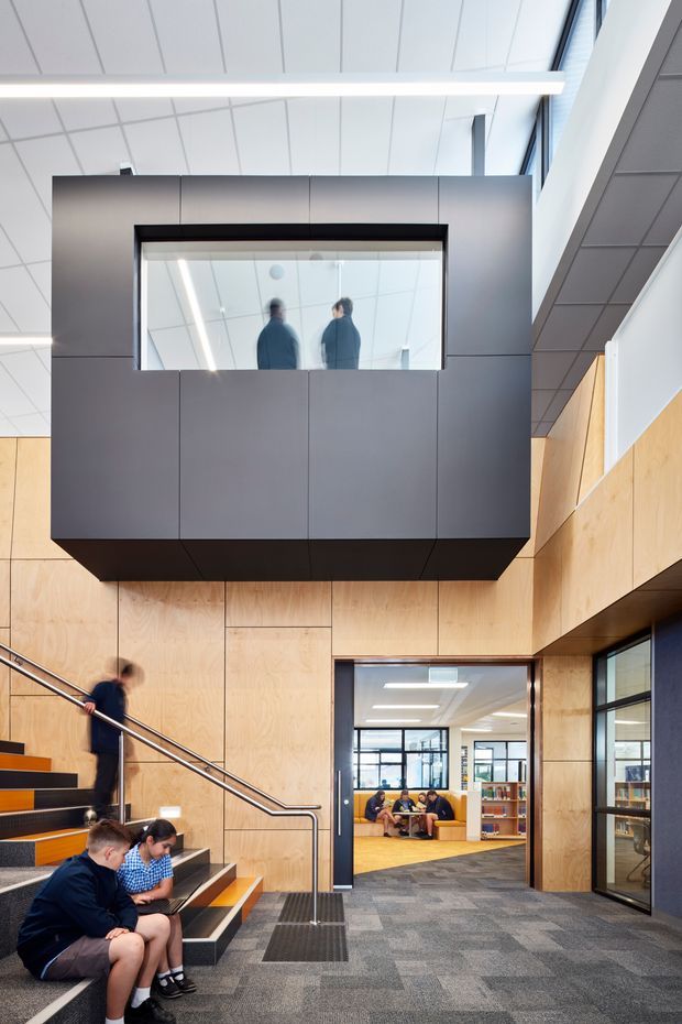 A bright contemporary multi-level educational or commercial interior featuring a stairway and landing with exposed timber wall panelling, dark grey suspended glazed structure, extensive internal glazing, and a carpeted floor. Students and seating are visible on the stairs and through the doorway, with a library or collaborative study area beyond. The space has a high ceiling, strong linear lighting, and a refined institutional design.