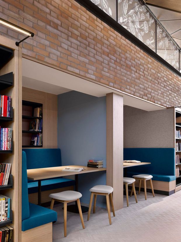 A contemporary library or study interior with built-in timber-framed study booths, deep blue upholstered bench seating, light timber desks, and white stools. Full-height bookshelves line the sides, while an exposed warm brick wall and glazed upper-level balustrade add an industrial architectural character. The space is softly lit and arranged for individual reading or study.