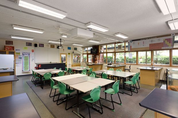 A bright, spacious classroom with rows of grouped rectangular tables and green student chairs. The room includes a whiteboard, wall-mounted educational displays, extensive windows, overhead fluorescent lighting, and a rear teaching or kitchenette-style counter area. The interior has a functional institutional fit-out with neutral flooring and walls, wood-framed windows, and colourful educational accents.