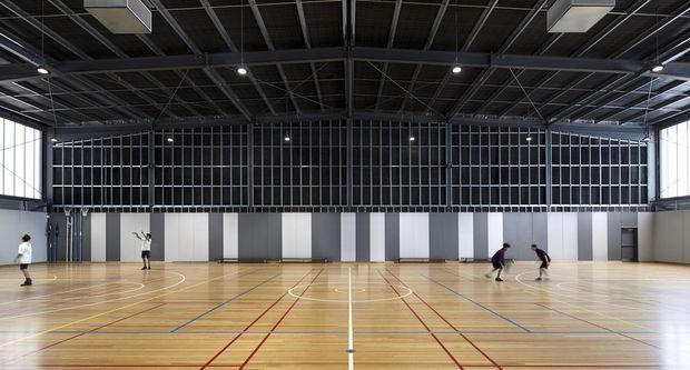 Large modern indoor sports hall with a polished timber multi-use court marked in white, red, blue and yellow lines. The space has a high exposed dark metal roof structure, suspended rectangular lights, tall glazed side walls, and a full-height dark and light panelled wall at the far end. Several people are using the court, emphasizing its substantial scale.