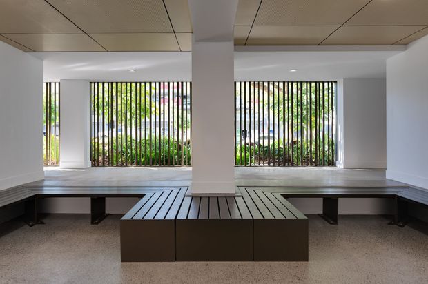 A minimalist interior space with built-in dark bench seating arranged around a central projecting column. Large windows with vertical black metal bars overlook dense greenery, while a light terrazzo-like floor and paneled ceiling create a clean, restrained architectural setting.