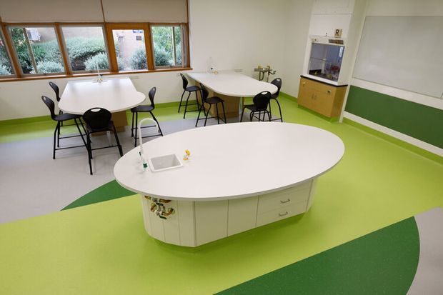 A bright classroom or educational activity room with white oval tables, black chairs, and a central curved island workstation or storage unit. The space features a vivid lime-green and dark-green patterned resilient floor, white walls, large timber-framed windows, and a small fixed kitchenette or storage cabinet at the rear. The room has a clean, functional institutional fit-out with abundant natural light.