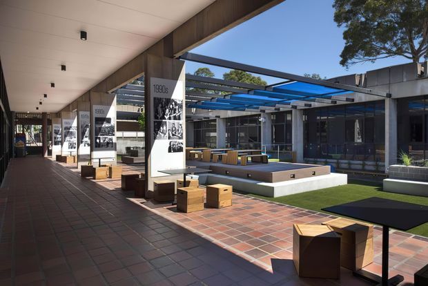 A covered outdoor courtyard or campus arcade with brick-paver flooring, dark timber-toned structural elements, and a central landscaped seating area. Modular timber seating and tables are arranged around a raised rectangular platform, while graphic display panels and glazed commercial or institutional spaces line the perimeter. Blue translucent overhead panels and exposed steel framing provide shade and create a contemporary, open-air gathering space.