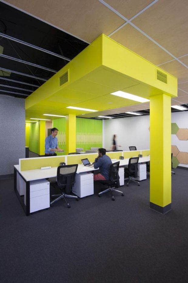 A brightly coloured open-plan commercial office with rows of white workstation desks and black ergonomic chairs. A vivid lime-yellow suspended ceiling canopy, structural columns, and partitions form the dominant feature, contrasting with dark carpet, exposed black ceiling services, white walls, and green vertical wall detailing. Hexagonal wall panels are visible in the background, with several occupants working at desks.