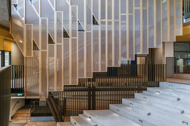 A contemporary multi-level interior circulation space featuring broad concrete stairs, black metal balustrades, and a dramatic sculptural stairwell screened by suspended translucent and timber-framed panels. The space appears to be a large institutional or commercial atrium with layered stairs, open landings, and strong vertical geometry.