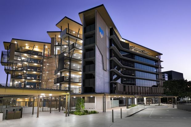 Twilight exterior view of a contemporary multi-storey commercial or apartment-style building. The complex features extensive glass curtain walls, projecting balconies, exposed vertical circulation elements, dark metal detailing, concrete structural walls, and warm interior lighting. A covered ground-level entry and driveway or forecourt are visible in front.