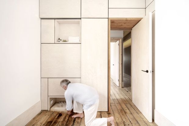 A minimalist interior hallway or entry area with extensive pale built-in cabinetry and open shelving, a warm reclaimed timber floor, and a timber-lined doorway leading to another room. A person is kneeling near a low recessed cabinet, while white doors and black hardware add contrast. The space is bright, clean, and highly integrated with concealed storage.