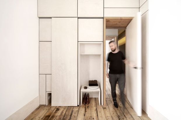 A compact minimalist entryway with full-height pale timber built-in cabinetry and a concealed doorway. The cabinetry includes a recessed open niche with a small seat, shoes, and accessories, while a person stands in the timber-lined passage beyond. The space has a reclaimed-looking timber floor, light neutral walls, clean geometric joinery, and a simple white door.