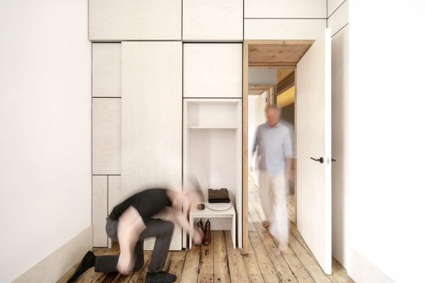 A narrow, minimalist interior hallway or entry area with full-height built-in pale timber cabinetry and open shelving along one wall. A white door opens into an adjoining space, where a blurred figure is visible. The room has distressed or textured light timber floorboards, white walls, simple black hardware, and a small built-in bench or shoe-storage nook. Motion-blurred people add a sense of activity while the fixed joinery dominates the composition.