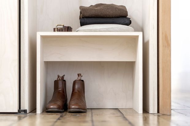 Close-up of a compact built-in entryway storage nook with an open light timber cubby bench or shelf. Folded clothing is stacked on top, while brown boots and a small suitcase occupy the lower compartments. Pale timber cabinetry and warm wood flooring create a simple, functional composition.