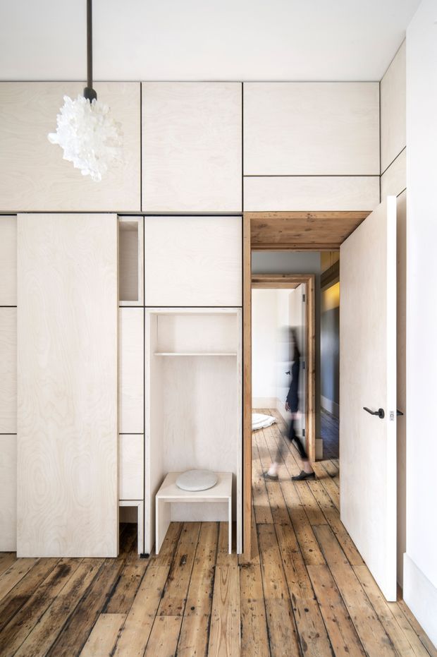 A bright, minimalist interior hallway or entry zone with extensive pale plywood built-in cabinetry and integrated open shelving. The space has exposed, weathered timber floorboards and a timber-lined passageway leading toward a bedroom or living area. A sculptural white pendant light hangs from the ceiling, while a white hinged door opens on the right. The design uses clean geometric lines, concealed storage, and a restrained natural palette.