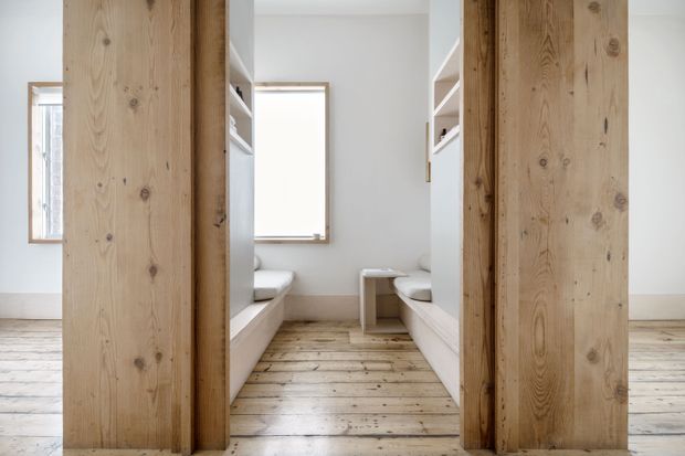 Bright, minimalist built-in seating nook viewed through a wide timber-framed opening. The room features white walls, pale exposed timber flooring, substantial natural timber posts and framing, built-in white benches on both sides, open shelving, and a central low table. Windows provide soft natural light.