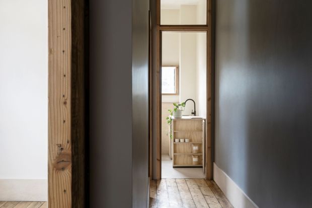 A narrow interior hallway with dark painted walls, pale timber flooring, and a timber-framed glazed doorway leading into a bright kitchen or utility area. The adjoining space contains a light timber cabinet or island with open shelving, a sink and curved black tap, and a potted plant. A structural timber post and white-floored room are visible at the left edge.