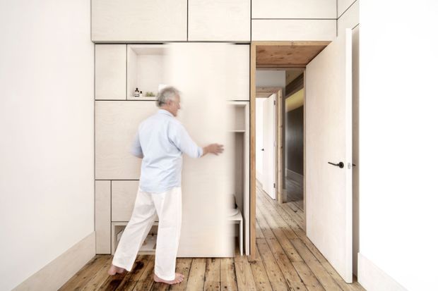 Interior view of a bright, minimalist hallway or flexible living space with pale full-height built-in cabinetry and a large sliding panel being opened. The room features a wide-plank natural timber floor, white walls and cabinetry, integrated shelving, and a timber-lined opening leading to a corridor with additional doors. A person is interacting with the joinery, demonstrating its movable or concealed-storage function.