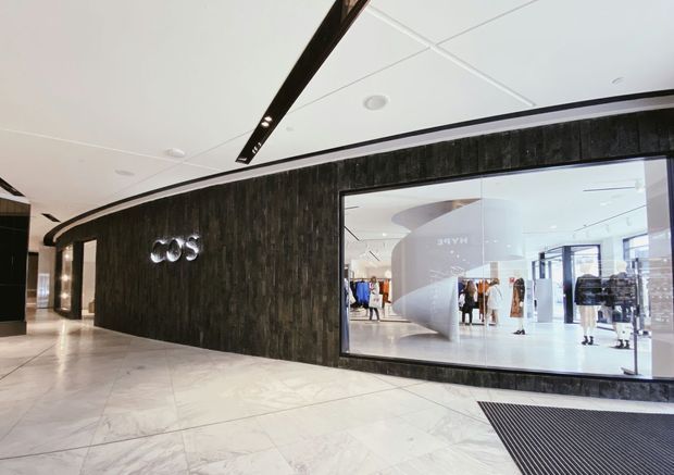 A polished contemporary retail storefront in a shopping centre, featuring a curved dark textured feature wall with illuminated signage, a large shopfront window displaying clothing and mannequins, pale stone-look tiled flooring, and recessed linear ceiling lighting.